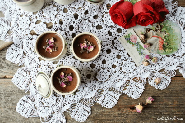 Three petit chocolate rose pots de crème topped with rose petals on a lace tablecloth with red roses.