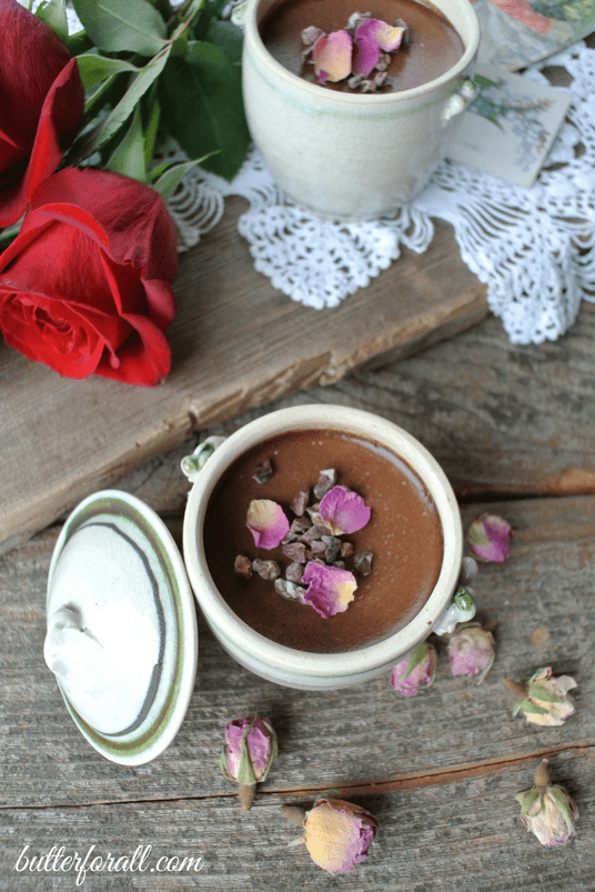 Petit chocolate rose pots de crème topped with rose petals on a wooden table with red roses.