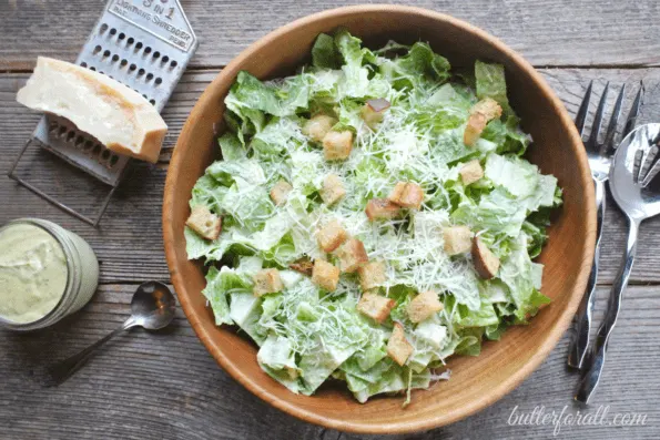 A big bowl of Ceasar salad with cheese and utensils on a wooden table.