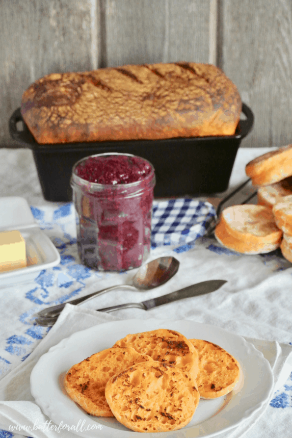 A breakfast spread with fresh sweet potato sourdough English muffins and a bread loaf with fresh butter and homemade jam. 