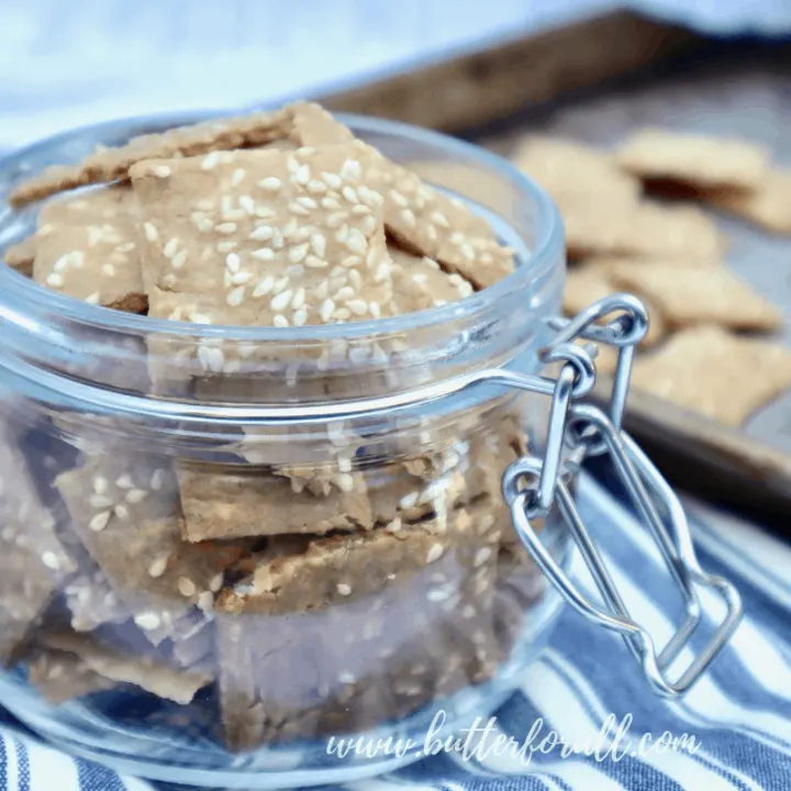 A Jar full of toasty brown sesame crackers with a baking sheet of crackers in the background.