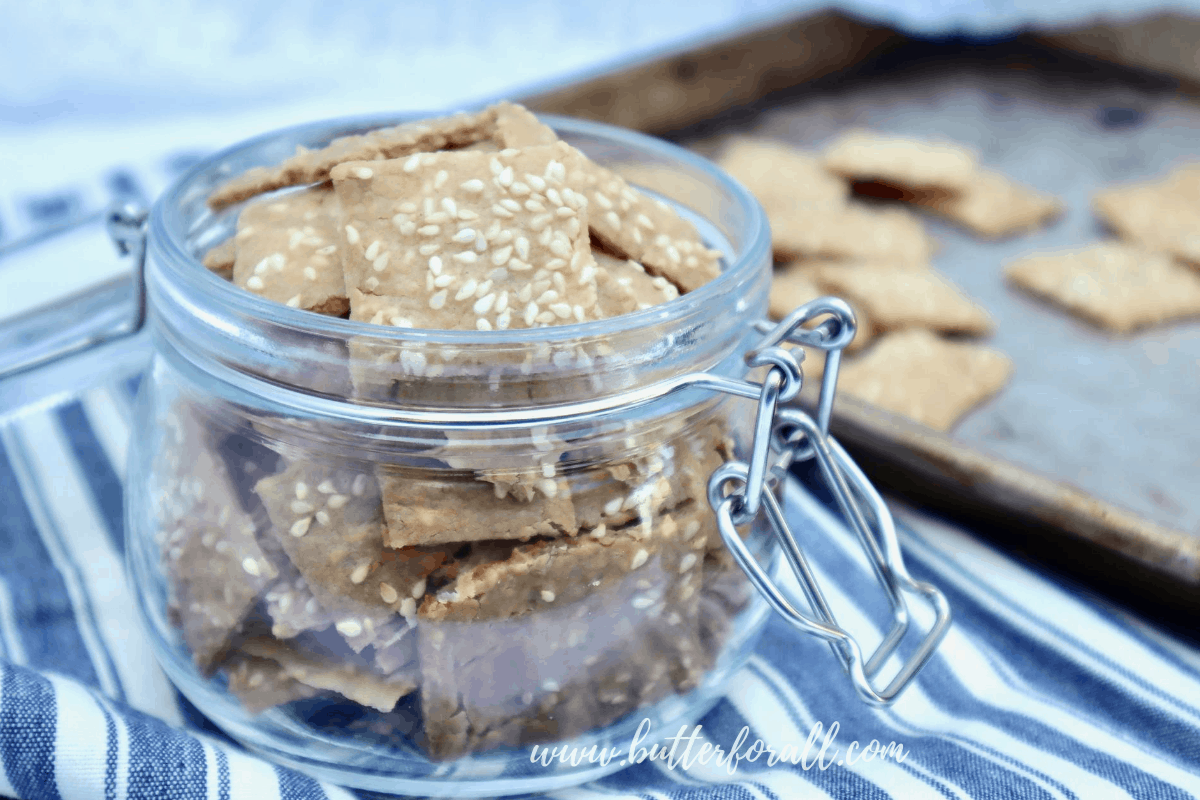 A Jar full of toasty brown sesame crackers with a baking sheet of crackers in the background.