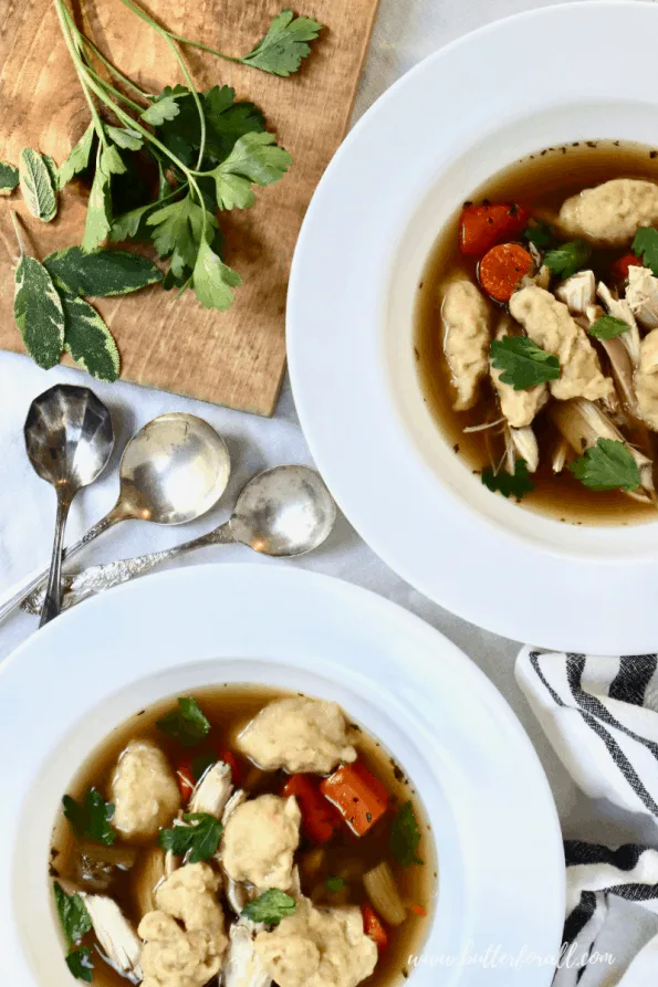 Bowls of clear chicken broth with cooked carrots, shredded chicken, sourdough dumplings, and fresh herbs.