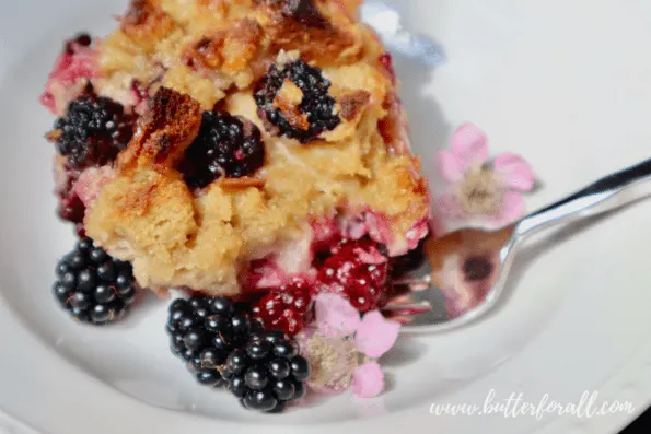 A white plate with a scoop of warm blackberry sourdough bread pudding adorned with fresh blackberries and blackberry flowers.