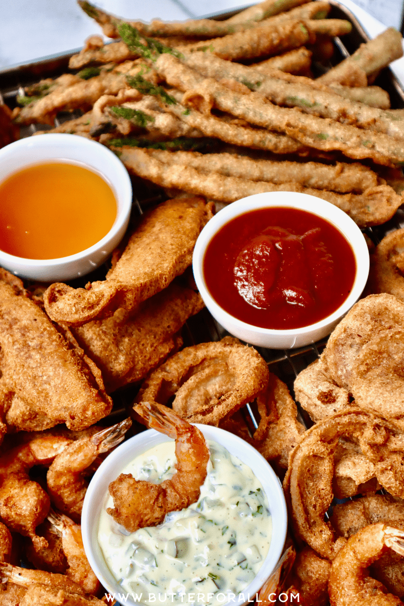 A platter of golden fried shrimp, chicken, onion rings, and asparagus spears with three dipping sauces.